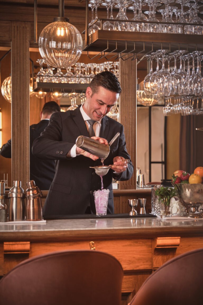Happy male bartender in black suit pouring cocktail from shaker through strainer in glass at counter in bar