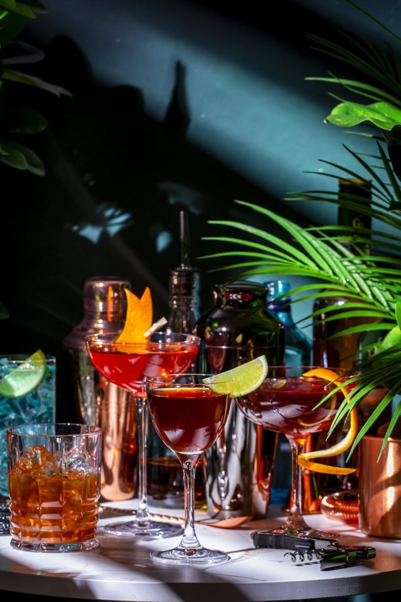 Summer alcoholic cocktail party with ready-made cocktails, bar tools and bottles of spirits on marble table with tropical plants. Green background, hard light and shadows. Shallow DOF